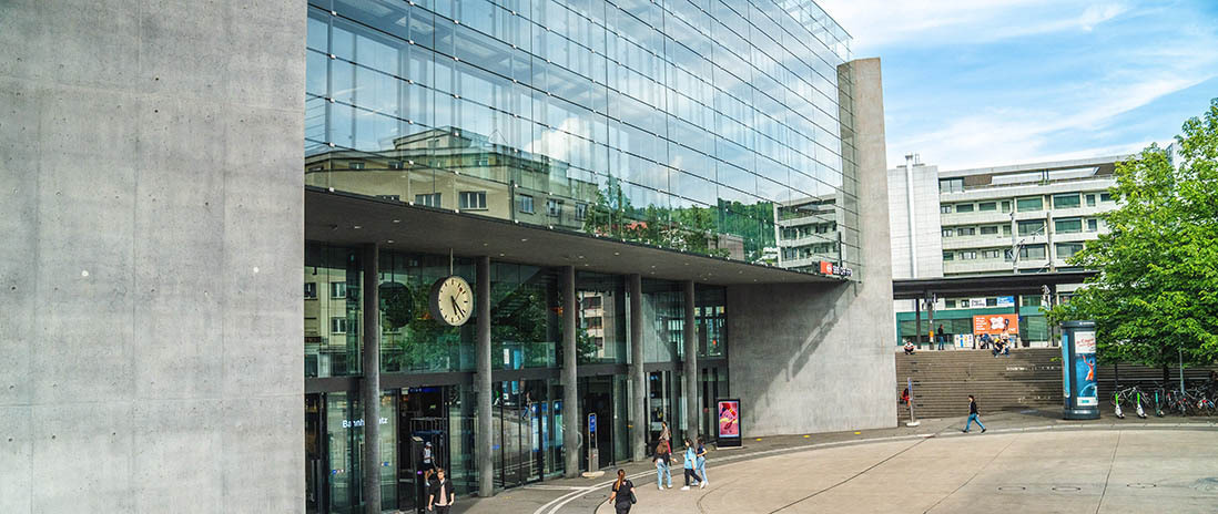 Moderner Eingangsbereich des Bahnhofs Zug mit Glasfassade und Platzgestaltung in der Nähe des Neubauprojekts Colin.