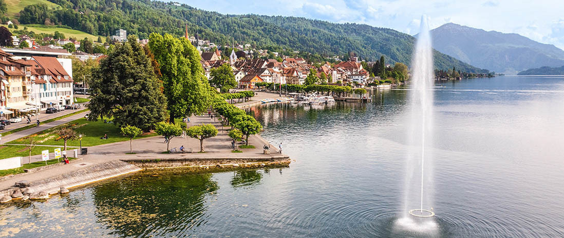 Blick auf die Zuger Seepromenade mit markantem Springbrunnen und historischer Altstadt im Hintergrund in der Nähe des Neubauprojekts Colin.